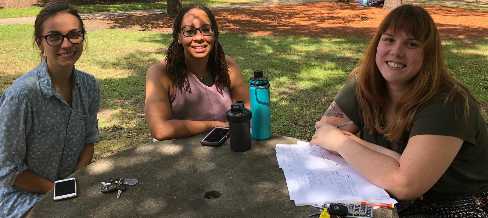 Three smiling, female, Caucasian students sitting at a round, grey stone table: the first has brown hair with blond highlights worn down and is wearing a black shirt and a black skirt with multi-colored polka dots and is pictured with a stack of paper under her crossed arms on the table with a ring of keys including yellow and white cards and a black cell phone; the second has brown hair worn down and black rimmed glasses with a lavendar pink shirt, she is pictured with a tall, teal and black water bottle, a shorter solid black water bottle, and her cell phone with a lavendar back and white edges; and the third has on a blue, collared button up shirt with white polka dots and tan pants, she is pictured with her car keys and a cell phone with white, horizontal bars on the upper and lower portion of the face.